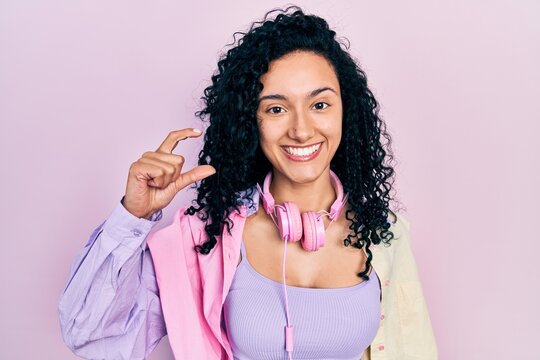 Young hispanic woman with curly hair wearing gym clothes and using headphones smiling and confident gesturing with hand doing small size sign with fingers looking and the camera. measure concept.