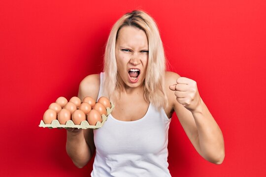 Beautiful caucasian blonde woman holding tray of fresh eggs annoyed and frustrated shouting with anger, yelling crazy with anger and hand raised