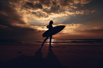 Silhouette of the surfer slim girl with surfboard in her hand on the sandy beach at sunset. Surfer...