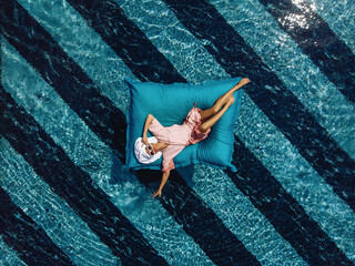 Top view of an adorable skinny girl in a bathrobe and with a towel on her head and sunglasses, floating in a pool on an inflatable mattress. Overflowing water. Summertime concept - obrazy, fototapety, plakaty