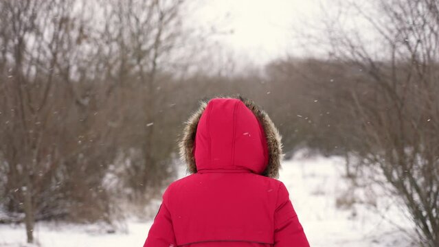 Anonymous woman wearing red hooded parka jacket standing under falling white snow in cold frosty winter forest landscape background. Slow motion 4k video footage of anonymous person enjoy solitude