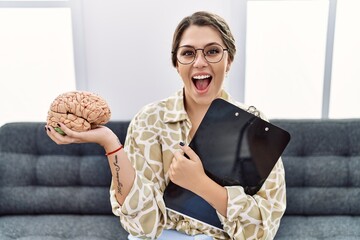Young hispanic woman holding brain working at psychology clinic celebrating crazy and amazed for...
