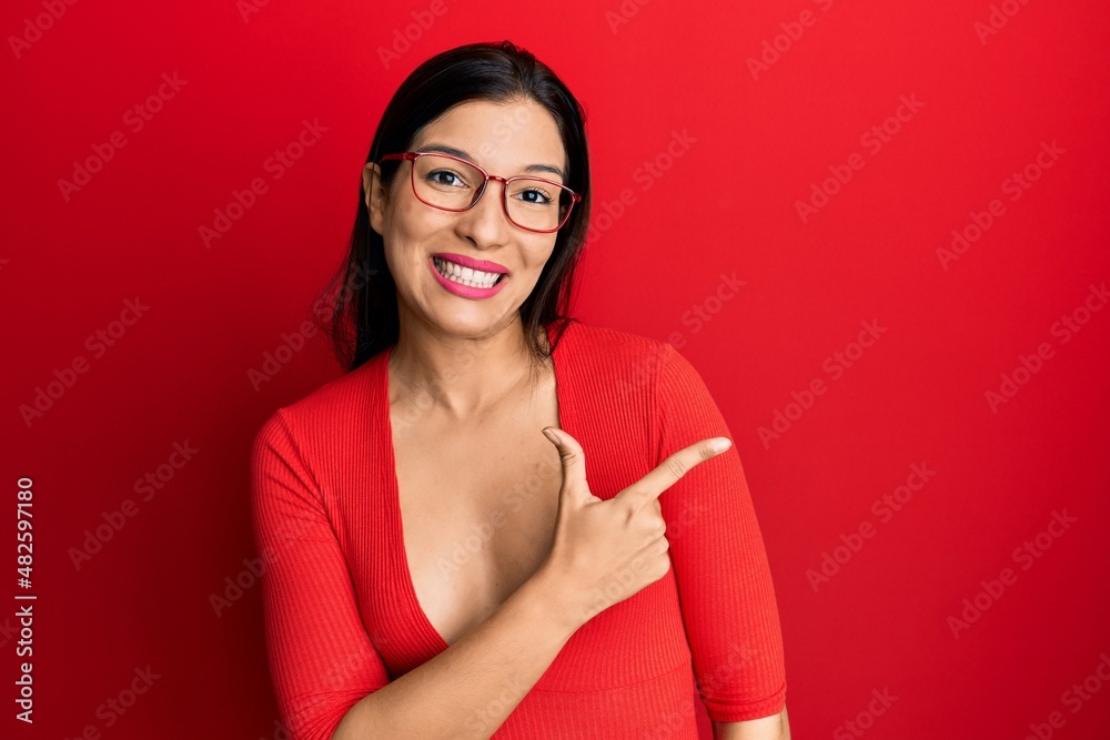 Sticker young latin woman wearing casual clothes and glasses cheerful with a smile of face pointing with han