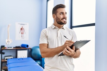 Young hispanic man wearing physiotherapist uniform writing on clipboard at clinic