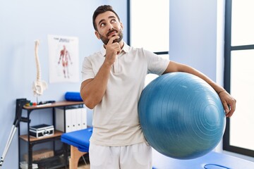 Handsome hispanic man holding pilates ball at rehabilitation clinic serious face thinking about question with hand on chin, thoughtful about confusing idea
