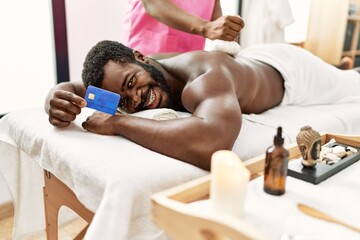 Obraz na płótnie Canvas Young african american man reciving herbal pouches thai massage and holding credit card at beauty center.