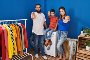Family of three at laundry room with open hand doing stop sign with serious and confident expression, defense gesture