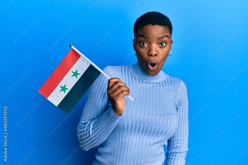 Sticker young african american woman holding syria flag scared and amazed with open mouth for surprise, disb