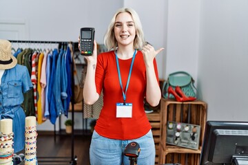 Naklejka na ściany i meble Young caucasian woman working as manager at retail boutique holding dataphone pointing thumb up to the side smiling happy with open mouth