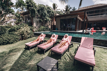 Three beautiful slender long-legged models lying on striped sunbeds sunbathing next to the pool and villa. Summer vacation concept - obrazy, fototapety, plakaty