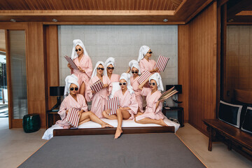 Spa treatments cute attractive ladies in bath clothes and sunglasses posing sitting on bed holding menu in hands indoors against wall background. Careless concept