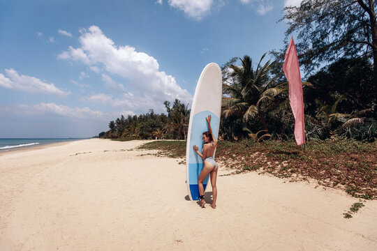 Charming hot slim woman in bikini posing on the white sand of a local beach holding a large surfboard with her hands on the background of the sea and nature. Hobby concept