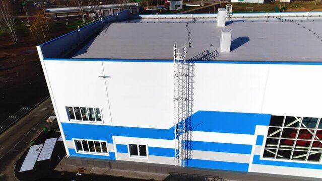 Large white and blue flat roofed warehouse building with windows and emergency ladder at plant area aerial view