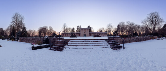 Bended panorama of snowy stairs on the cemetery at sunset time with clear sky and bald trees