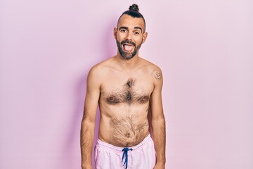 Fototapeta premium Young hispanic man shirtless wearing swimsuit sticking tongue out happy with funny expression. emotion concept.