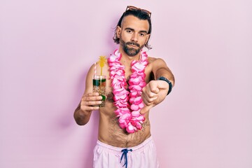 Young hispanic man wearing swimsuit and hawaiian lei drinking tropical cocktail looking unhappy and...
