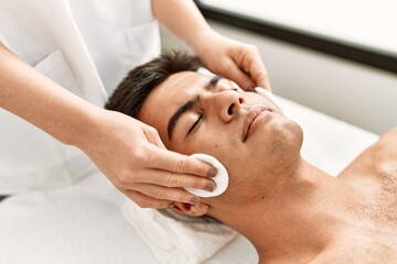 Young hispanic man relaxed having facial treatment cleaning face with cotton disk at beauty center
