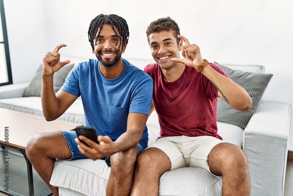 Sticker Young hispanic men using smartphone sitting on the sofa at home smiling and confident gesturing with hand doing small size sign with fingers looking and the camera. measure concept.