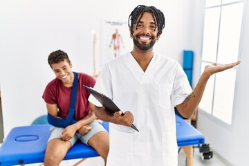 Obraz premium Young hispanic man working at pain recovery clinic with a man with broken arm smiling cheerful presenting and pointing with palm of hand looking at the camera.