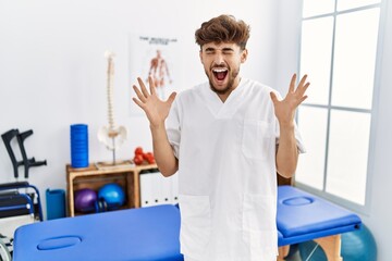 Young arab man working at pain recovery clinic celebrating mad and crazy for success with arms raised and closed eyes screaming excited. winner concept