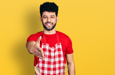 Young arab man with beard wearing cook apron smiling friendly offering handshake as greeting and welcoming. successful business.