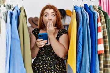 Young redhead woman searching clothes on clothing rack using smartphone covering mouth with hand, shocked and afraid for mistake. surprised expression