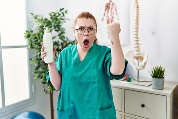 Young redhead physiotherapist woman holding massage body lotion annoyed and frustrated shouting...