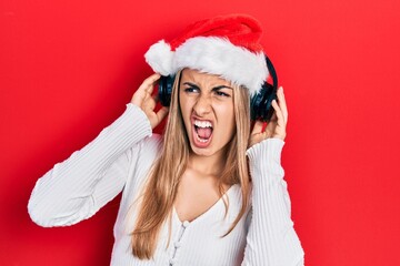 Beautiful hispanic woman wearing christmas hat and headphones angry and mad screaming frustrated and furious, shouting with anger. rage and aggressive concept.