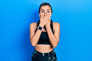 Fototapeta na wymiar Beautiful hispanic woman wearing gym clothes and using headphones shocked covering mouth with hands for mistake. secret concept.