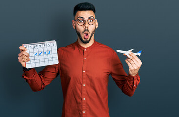 Young hispanic man with beard holding plane toy and travel calendar afraid and shocked with...