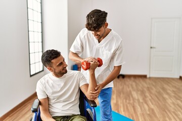 Two hispanic men physiotherapist and patient sitting on wheelchair having rehab session using dumbbell at clinic