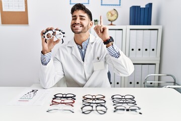 Young optician man holding optometry glasses pointing finger up with successful idea. exited and...