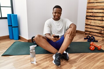 Young african man sitting on training mat at the gym using smartphone smiling looking to the side and staring away thinking.