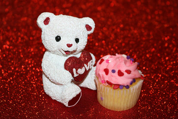 White teddy bear holding red glitter heart sitting beside a cupcake, on a red glitter background.