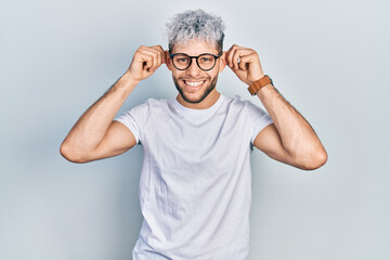 Young hispanic man with modern dyed hair wearing white t shirt and glasses smiling pulling ears...