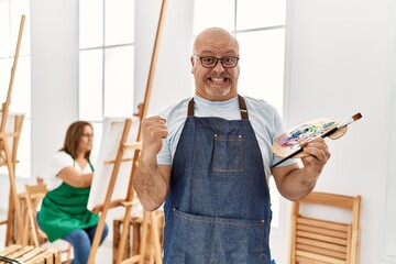 Middle age hispanic man at art studio screaming proud, celebrating victory and success very excited with raised arms