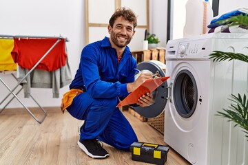 Young hispanic man wearing handyman uniform repairing washing machine at laundry room