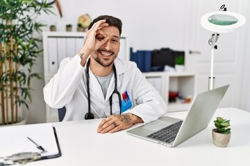 Young doctor working at the clinic using computer laptop doing ok gesture with hand smiling, eye looking through fingers with happy face.