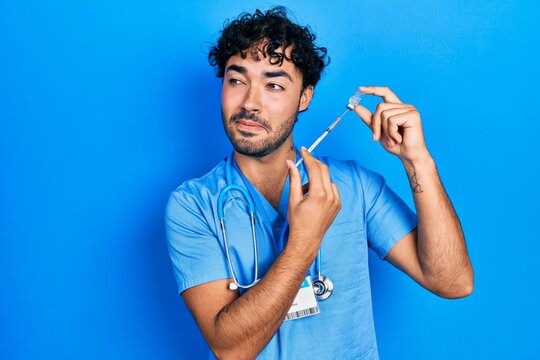 Young hispanic man wearing nurse uniform holding vaccine smiling looking to the side and staring away thinking.