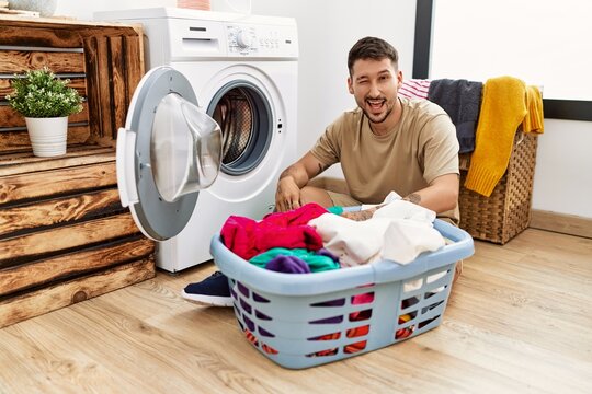 Young handsome man putting dirty laundry into washing machine winking looking at the camera with sexy expression, cheerful and happy face.