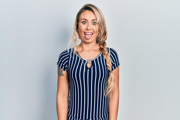 Beautiful young blonde woman wearing casual striped dress sticking tongue out happy with funny expression. emotion concept.