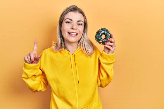 Beautiful caucasian woman holding donut smiling with an idea or question pointing finger with happy face, number one