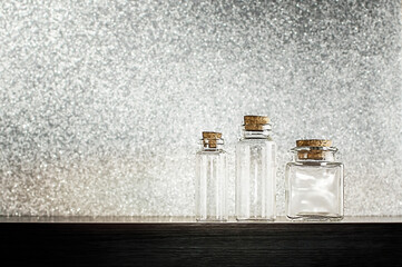 Obraz na płótnie Canvas Small empty glass bottles on wooden shelf against gray glitter background. Front view on clear small empty glass vials with cork. Copy space. Selective focus. 