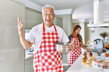 Middle age hispanic couple wearing apron cooking homemade pastry celebrating victory with happy smile and winner expression with raised hands