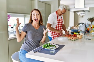 Middle age hispanic couple eating healthy salad at home celebrating victory with happy smile and winner expression with raised hands