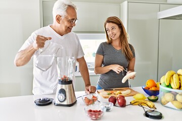 Middle age hispanic couple pouring water on mixer machine cooking smoothie at the kitchen.