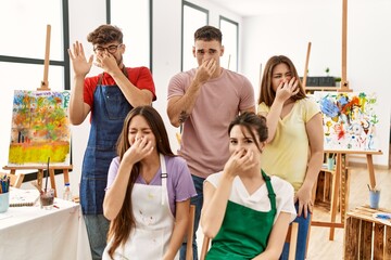 Group of five hispanic artists at art studio smelling something stinky and disgusting, intolerable smell, holding breath with fingers on nose. bad smell