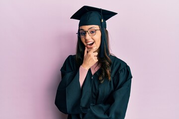 Young hispanic woman wearing graduation cap and ceremony robe looking confident at the camera...