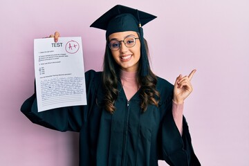 Young hispanic woman wearing graduation uniform holding passed exam smiling happy pointing with hand and finger to the side