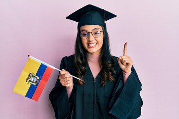 Young hispanic woman wearing graduation uniform holding ecuador flag smiling with an idea or question pointing finger with happy face, number one